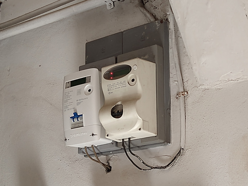 Two power meters - an older one and a newer one - placed on a wall. Both have wires going back to the distribution board clearly visible.