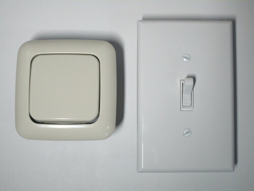 Front view of a European light switch, on the left, and an American one on the right. The American light switch has a large cover plate, but only a small part of it is used for the switch itself.